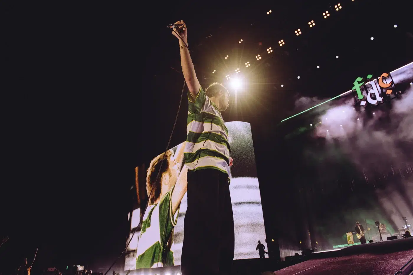 Grian Chatten of Fontaines D.C. raising an arm on stage at Reading Festival in the Cwmbran Celtic Mogwai shirt, the shirt shown again on the screen behind him